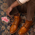 Pair of tan leather cap toe boots with brown laces, worn by a person sitting on a patterned rug next to two playing cards.