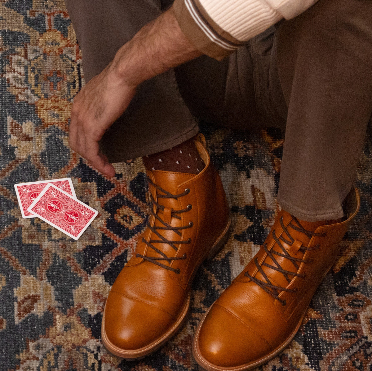 Pair of tan leather cap toe boots with brown laces, worn by a person sitting on a patterned rug next to two playing cards.