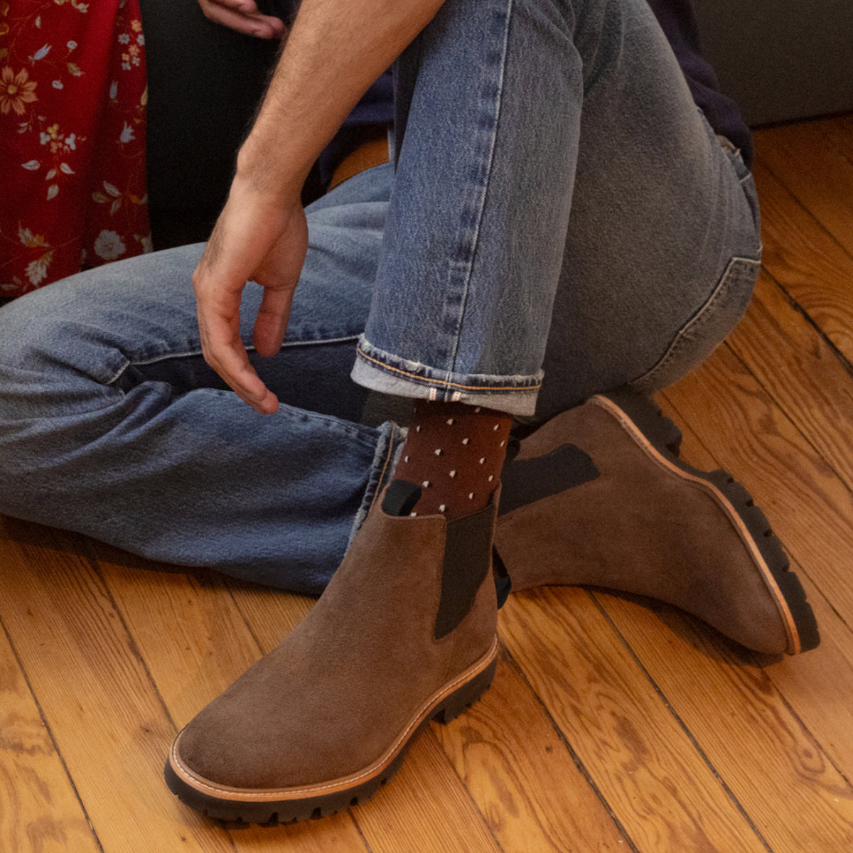 A person wearing brown suede Chelsea boots and blue denim jeans sits on a wooden floor.