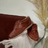 Two brandy brown leather woven huarache oxford shoes resting on a white patterned blanket next to a glass vase.