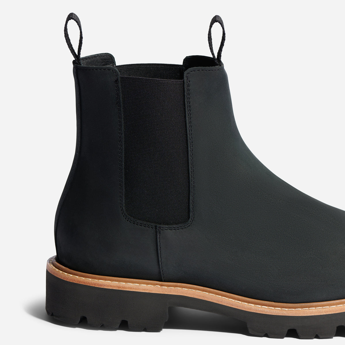 Side view of a black leather Chelsea boot with elastic side panels, pull tabs, and a lugged sole with tan welt stitching.