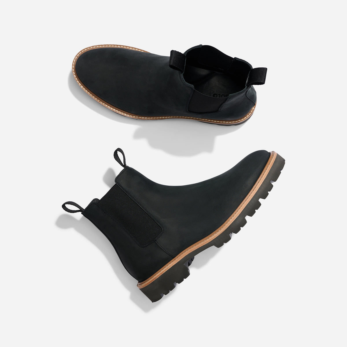 Pair of black leather Chelsea boots with tan midsoles and lugged outsoles, shown from side and top views on a white surface.