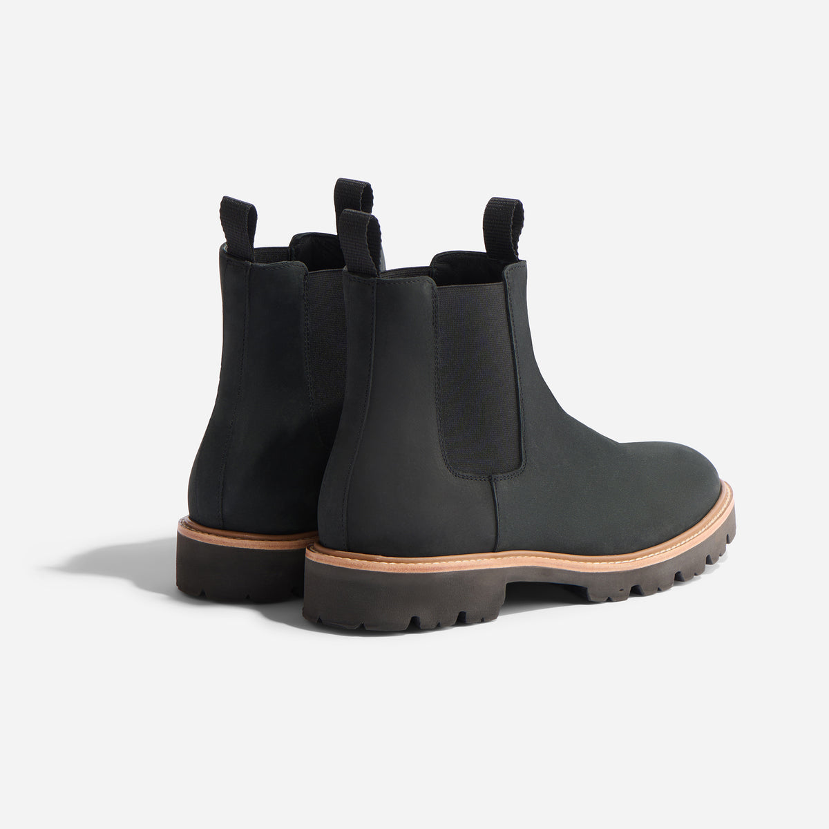 Pair of black leather Chelsea boots with tan midsoles, lugged outsoles, and fabric pull tabs on a white background.