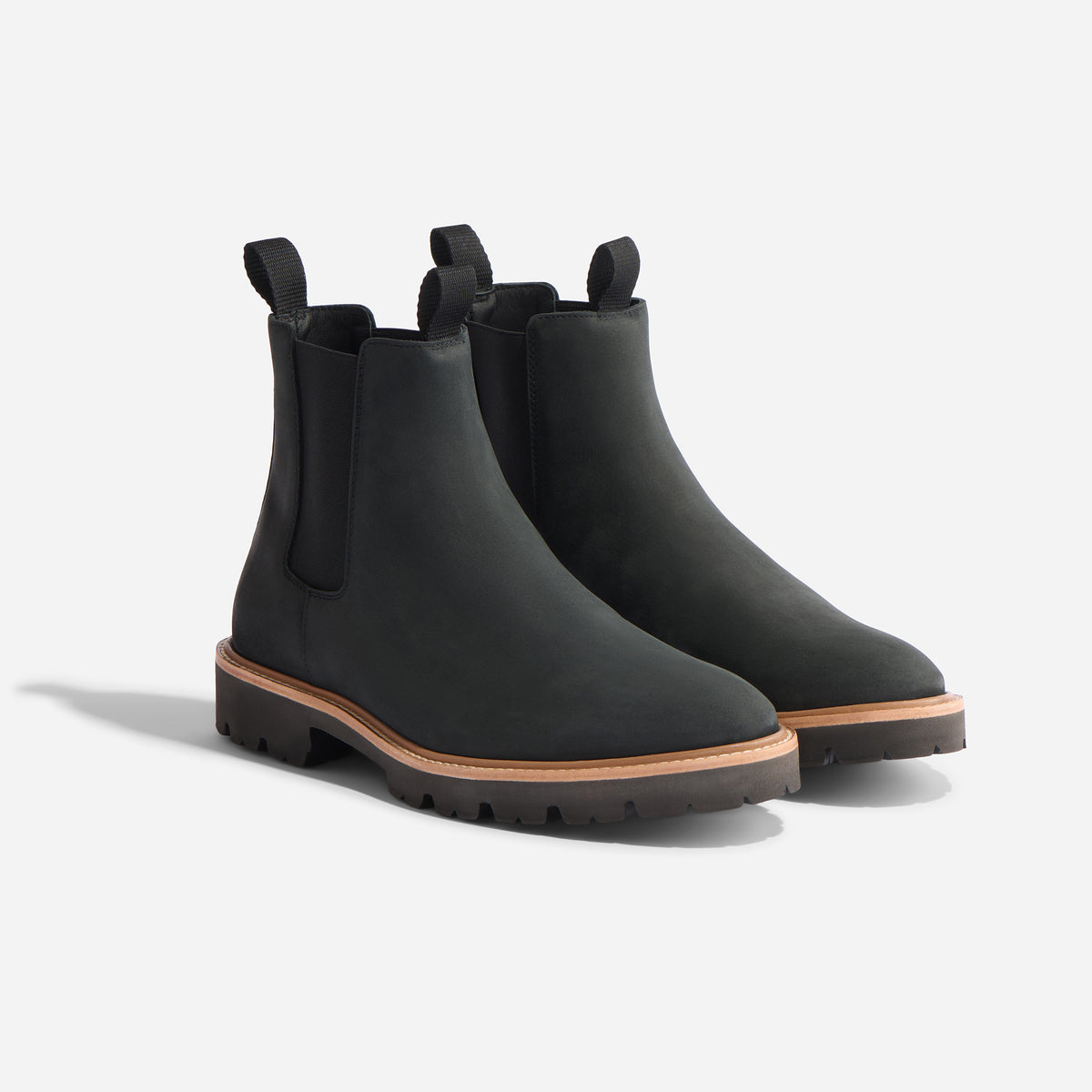 Pair of black matte Chelsea boots with elastic side panels, pull tabs, and tan welt stitching on lugged rubber soles.