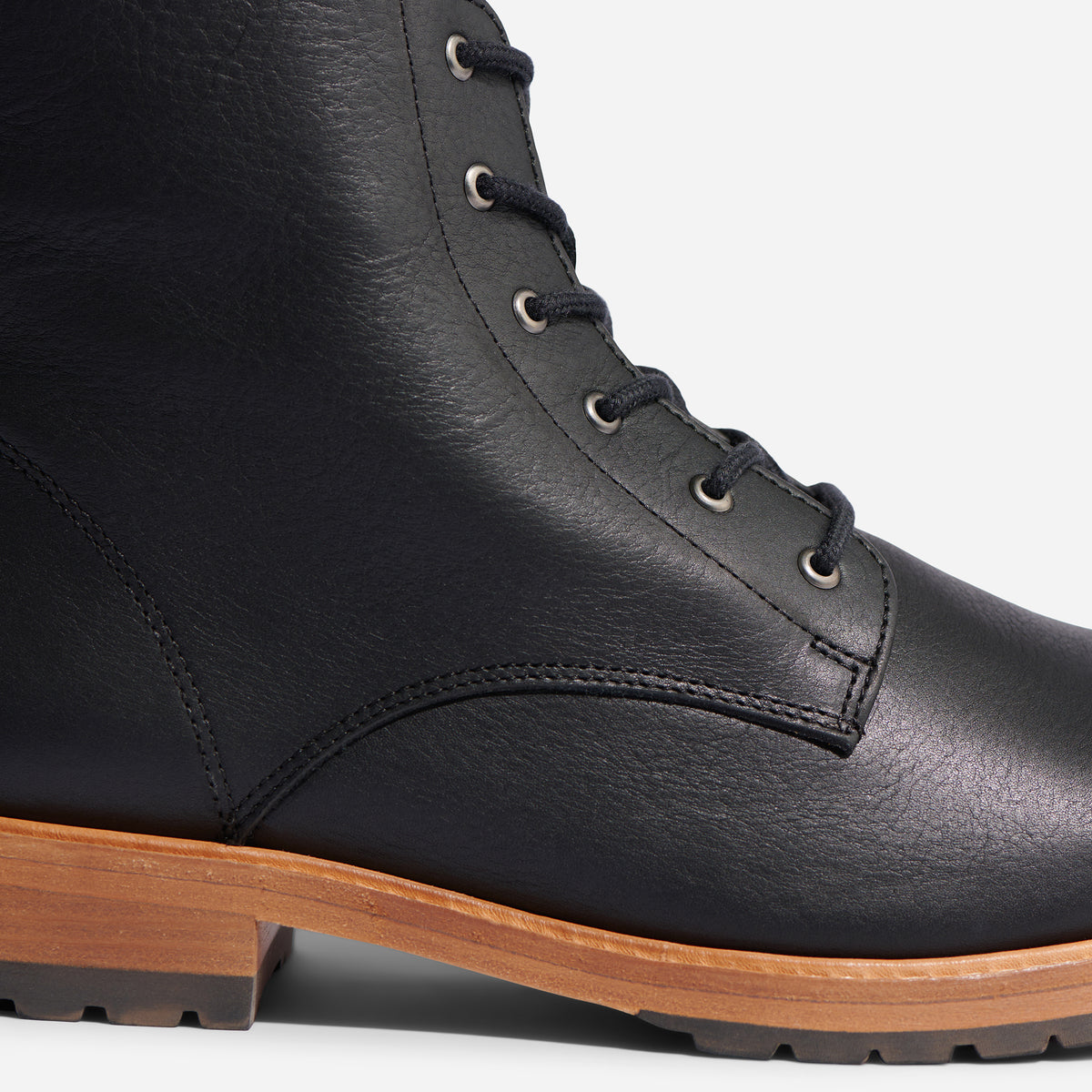 Close-up of a black leather boot with metal eyelets, black laces, and a light brown lug sole.