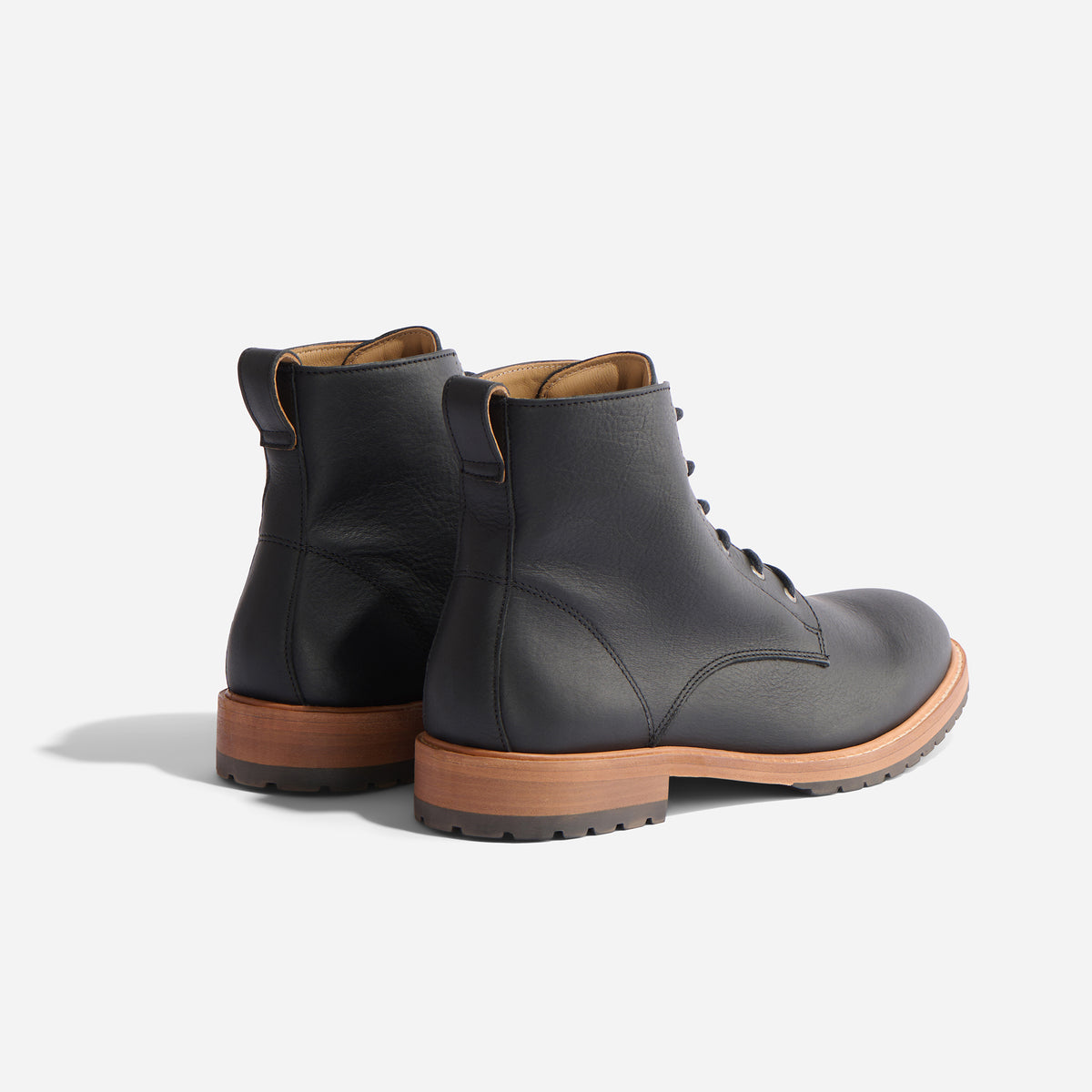 Pair of black leather lace-up boots with tan soles and pull tabs, shown from a rear three-quarter angle on white.