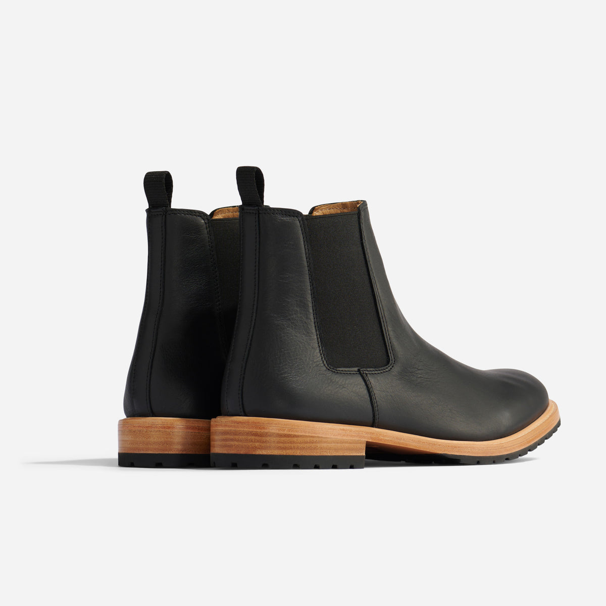 Pair of black leather Chelsea boots with tan midsoles and black lug outsoles, shown from a rear three-quarter angle.