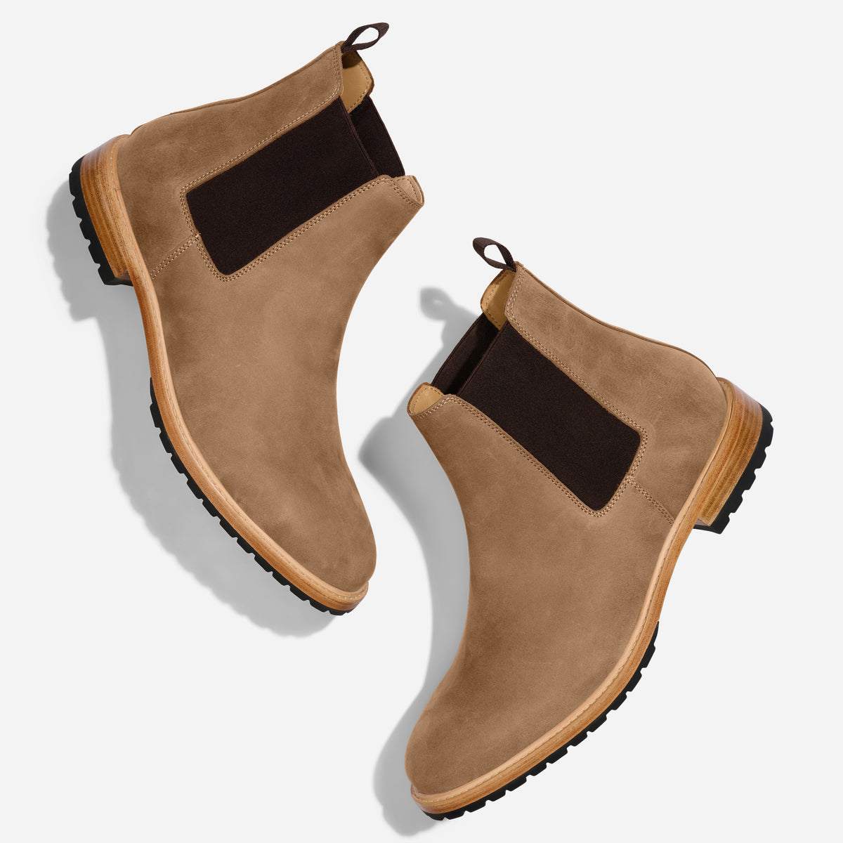 Pair of tobacco brown suede Chelsea boots with dark elastic side panels and lugged rubber soles on a white background.