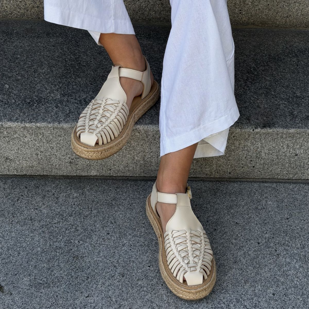 Pair of bone white woven leather T-strap sandals with jute espadrille soles worn on feet against stone steps.