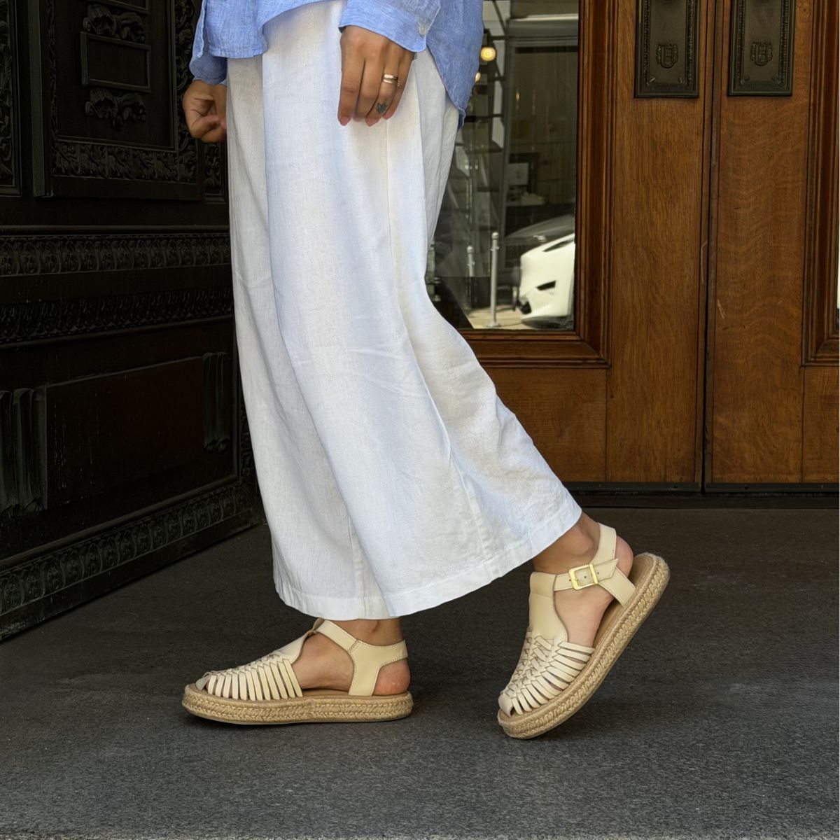 Pair of bone-colored leather huarache T-strap espadrille sandals with woven toes and jute soles worn with white pants.