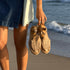 A pair of almond leather huarache T-strap espadrille sandals held by a person standing on a sandy beach.