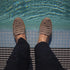 Pair of greige woven leather Huarache sandals on feet, standing at the edge of a blue tiled pool.
