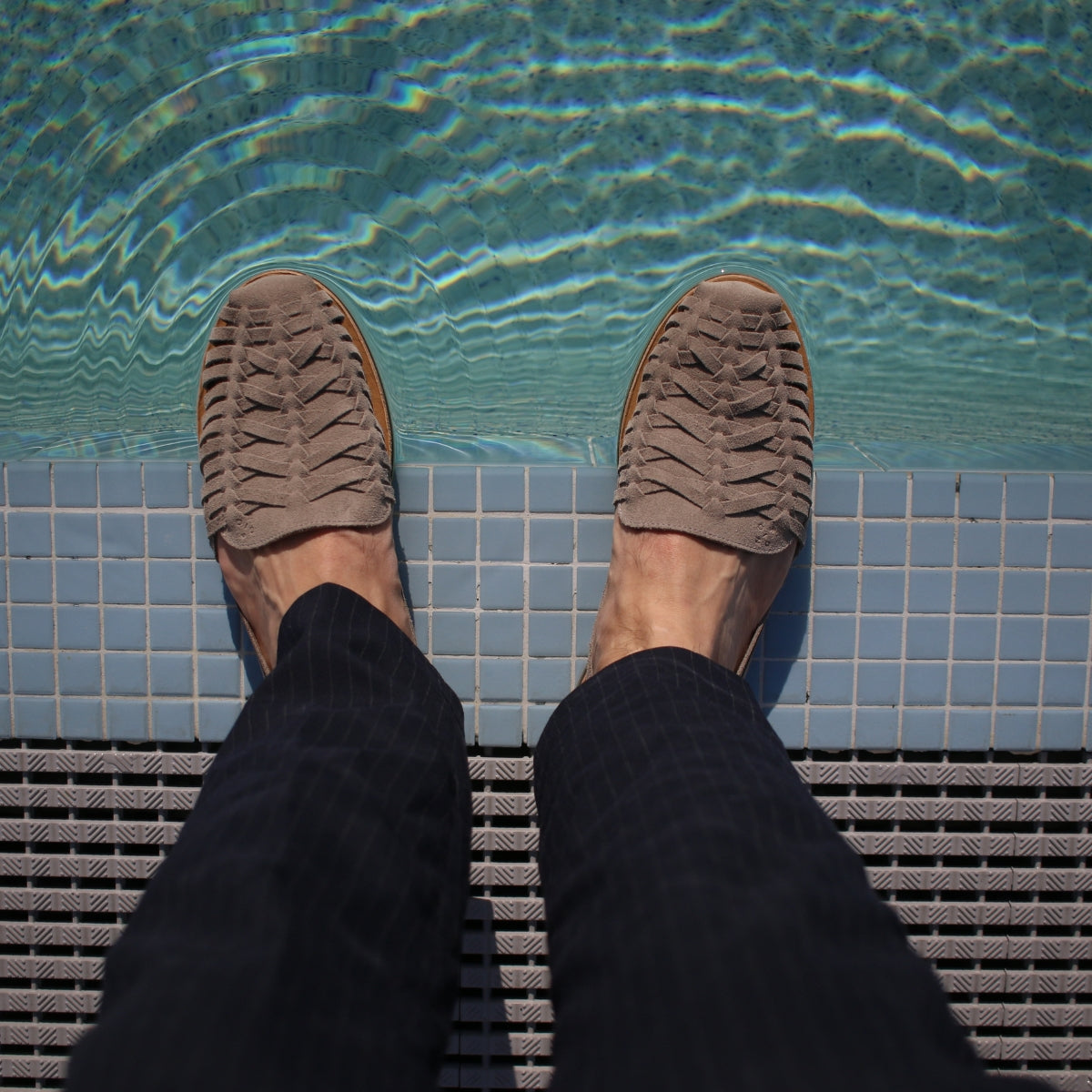 Pair of greige woven leather Huarache sandals on feet, standing at the edge of a blue tiled pool.