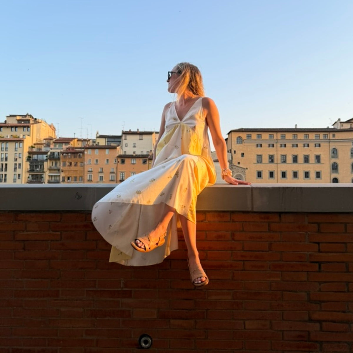 Woman in a white dress wearing tan leather Suri Sandals while sitting on a brick wall with a city skyline background.