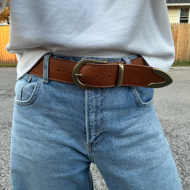 Brown textured leather belt with a gold-tone horseshoe buckle and metal tip worn on blue denim jeans.