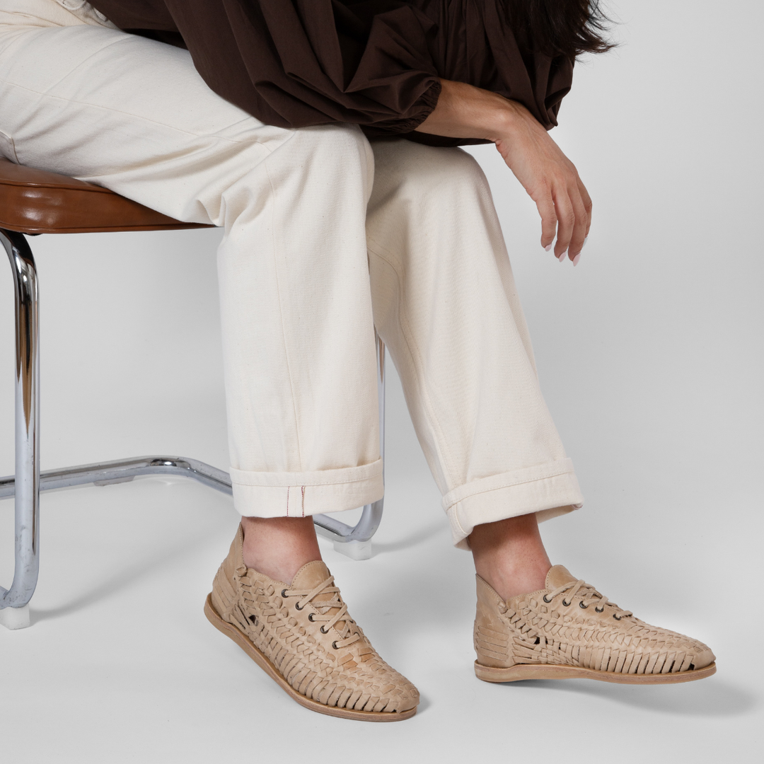 Pair of almond tan woven leather huarache booties with laces worn with white pants.