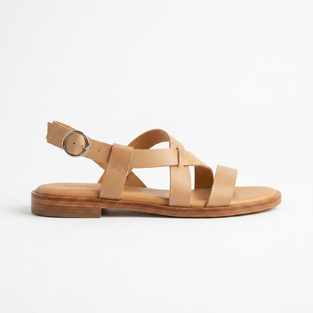 Single tan leather Suri Sandal with crisscross straps and a silver buckle on a flat wood-tone sole.