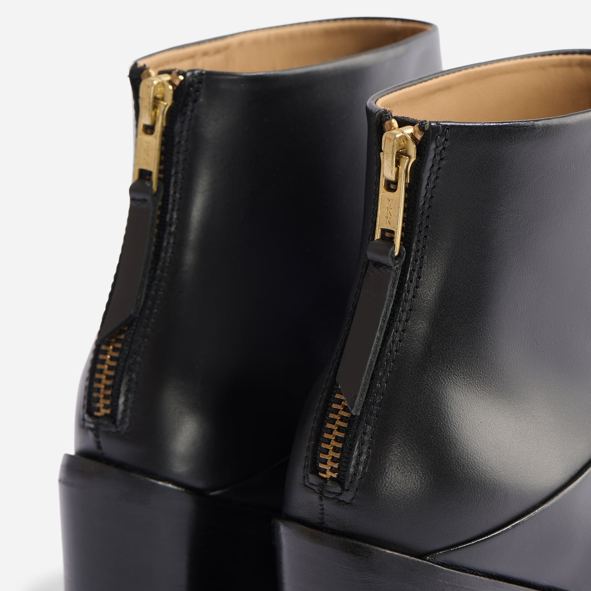 Rear view of two black leather Dari Boot 2.0 shoes featuring gold-tone metal zippers with leather pull tabs.