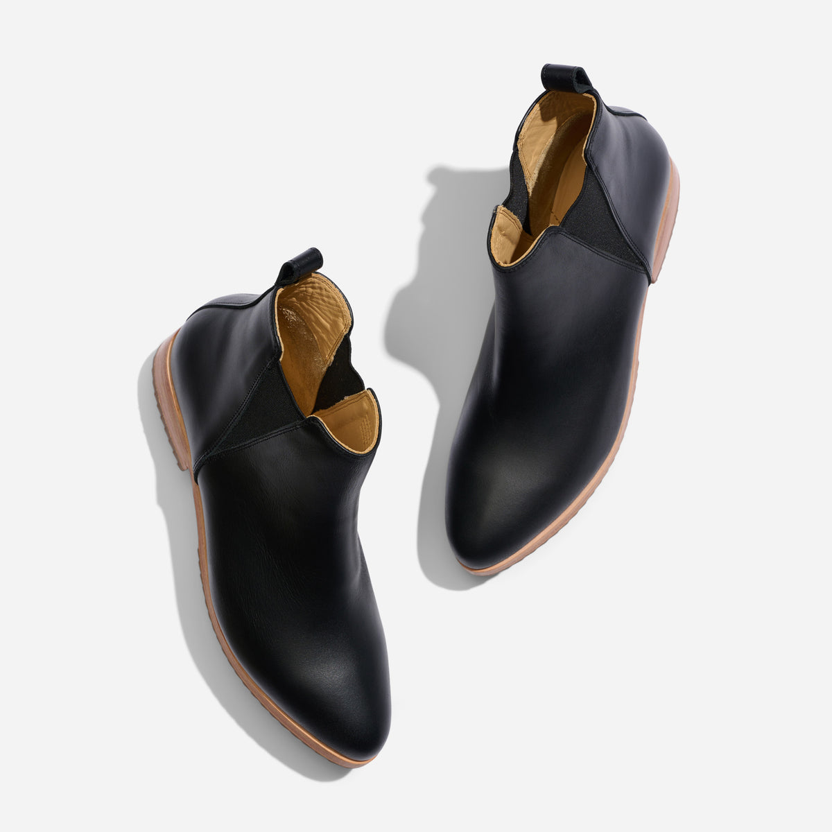 Pair of black leather Chelsea boots with tan soles and elastic side panels, viewed from above on a white background.