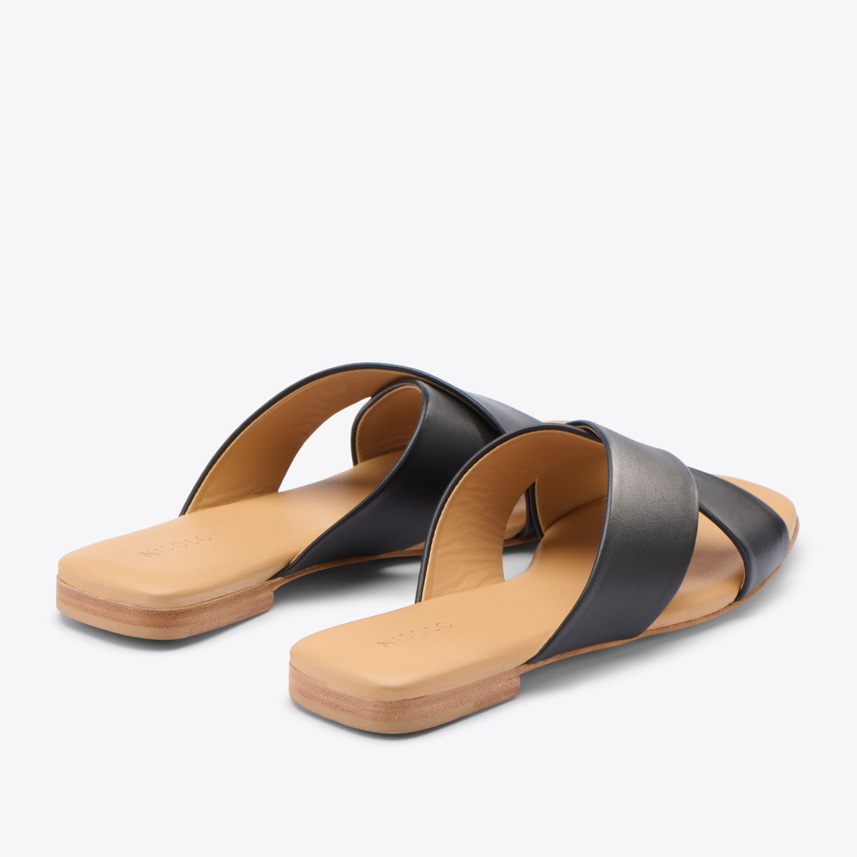 Pair of black leather slide sandals with tan footbeds and crisscross straps, featuring the NISOLO logo on the heel.