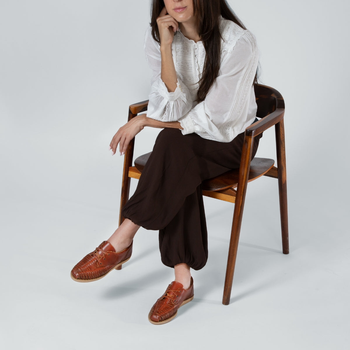 A woman sitting in a wooden chair wearing brandy-colored woven leather Huarache Oxford shoes and brown trousers.