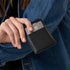 A hand holding a black pebbled leather Nico Card Case containing a silver metal credit card against a denim jacket.