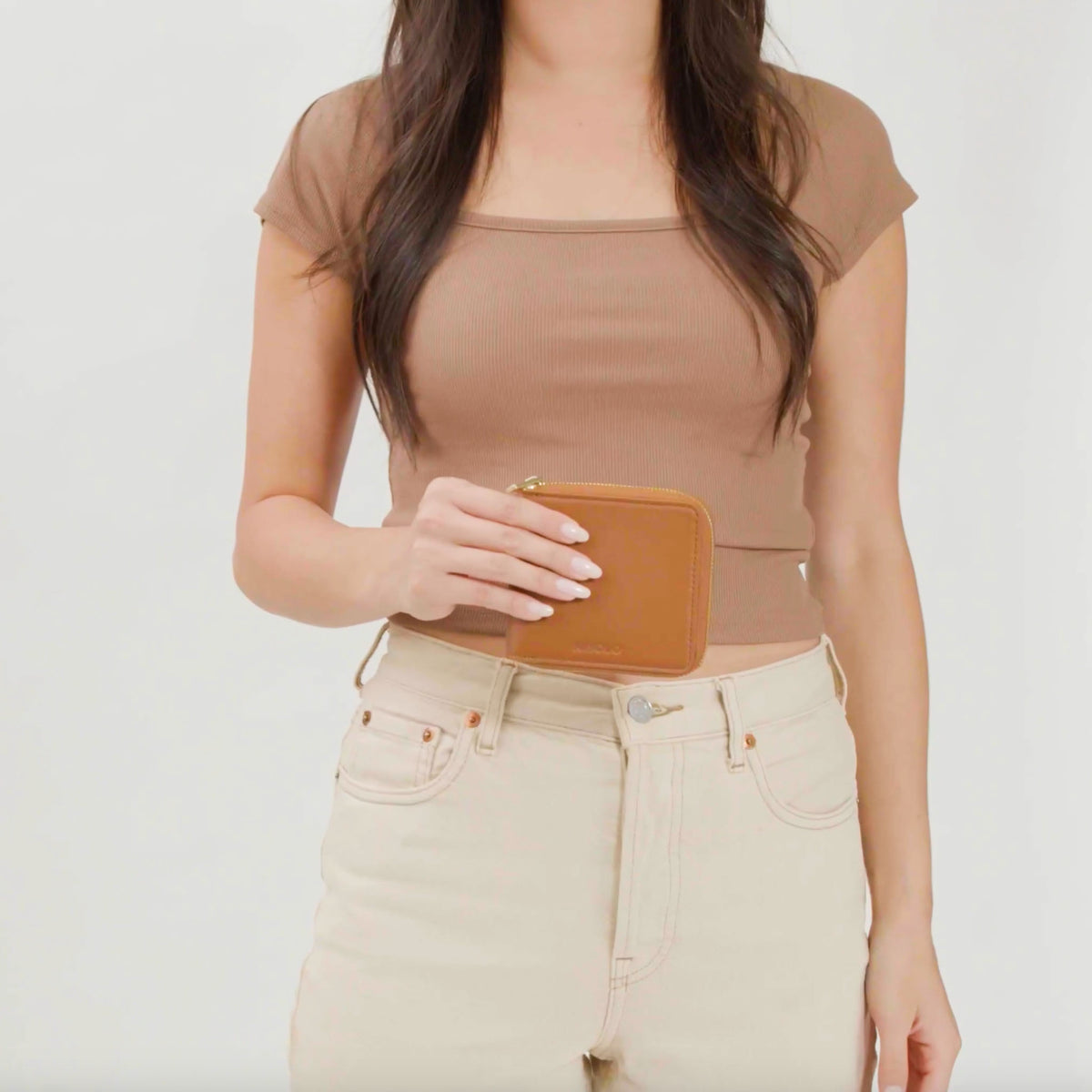 A woman holds a square caramel leather Remi Zip Wallet with a gold zipper against a white background.
