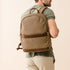 Tan fabric backpack with brown leather trim and top handle worn by a person. The brand name NISOLO is printed on the bottom.