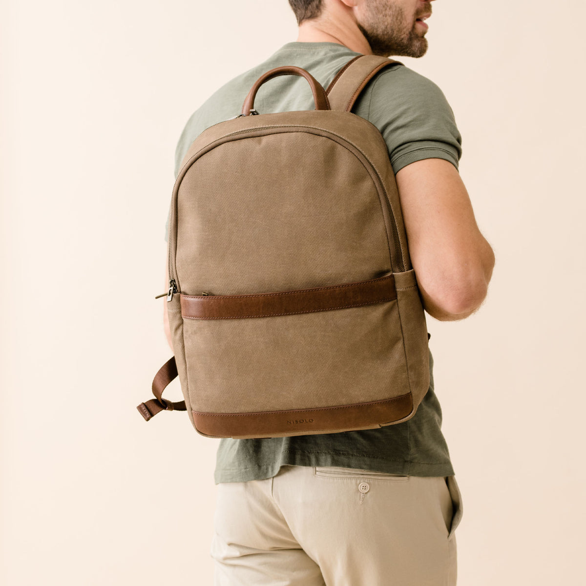 Tan fabric backpack with brown leather trim and top handle worn by a person. The brand name NISOLO is printed on the bottom.