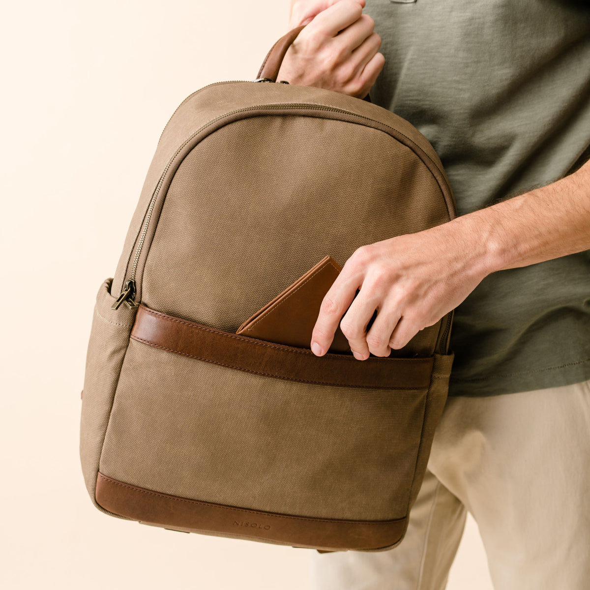 Brown waxed canvas NISOLO backpack with leather trim being held by a person placing a wallet in the front pocket.