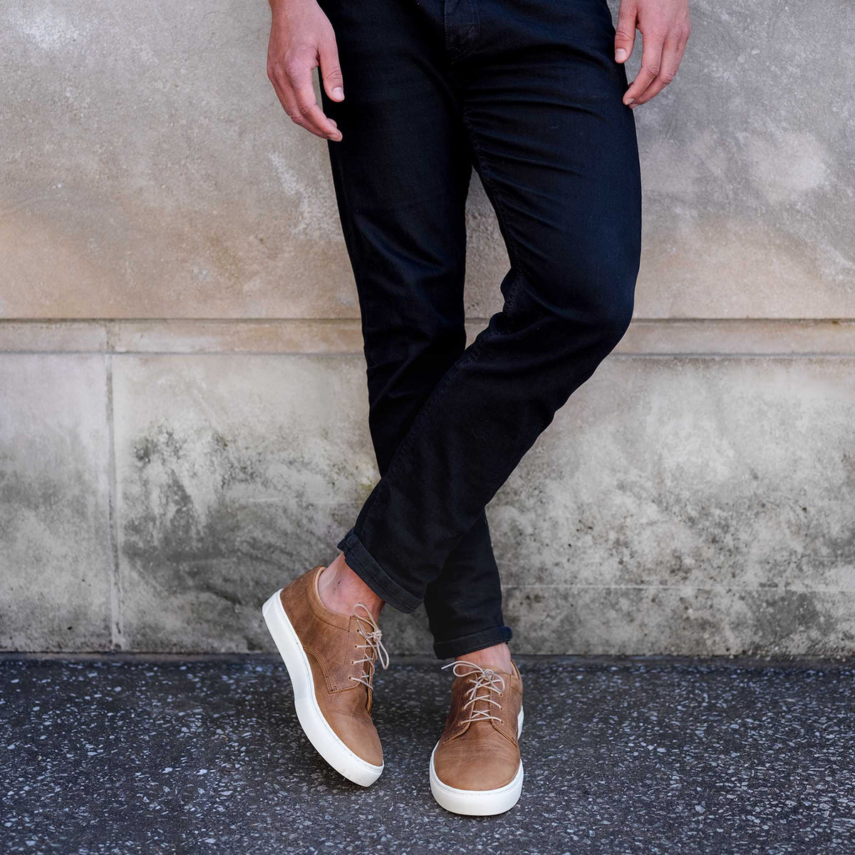 Pair of brown leather sneakers with white soles and tan laces worn with black pants against a stone wall.