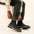 Pair of black leather Chelsea boots with lug soles and elastic side panels worn by a person sitting on a wooden box.