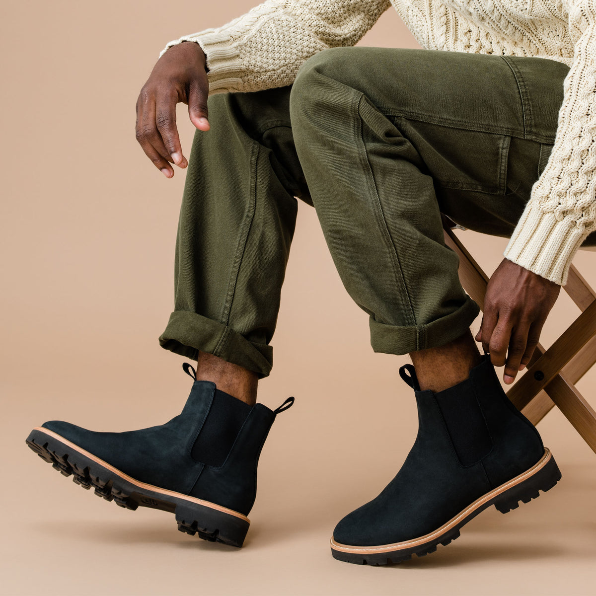 Pair of black leather Chelsea boots with lug soles worn by a person in green pants and a cream knit sweater.