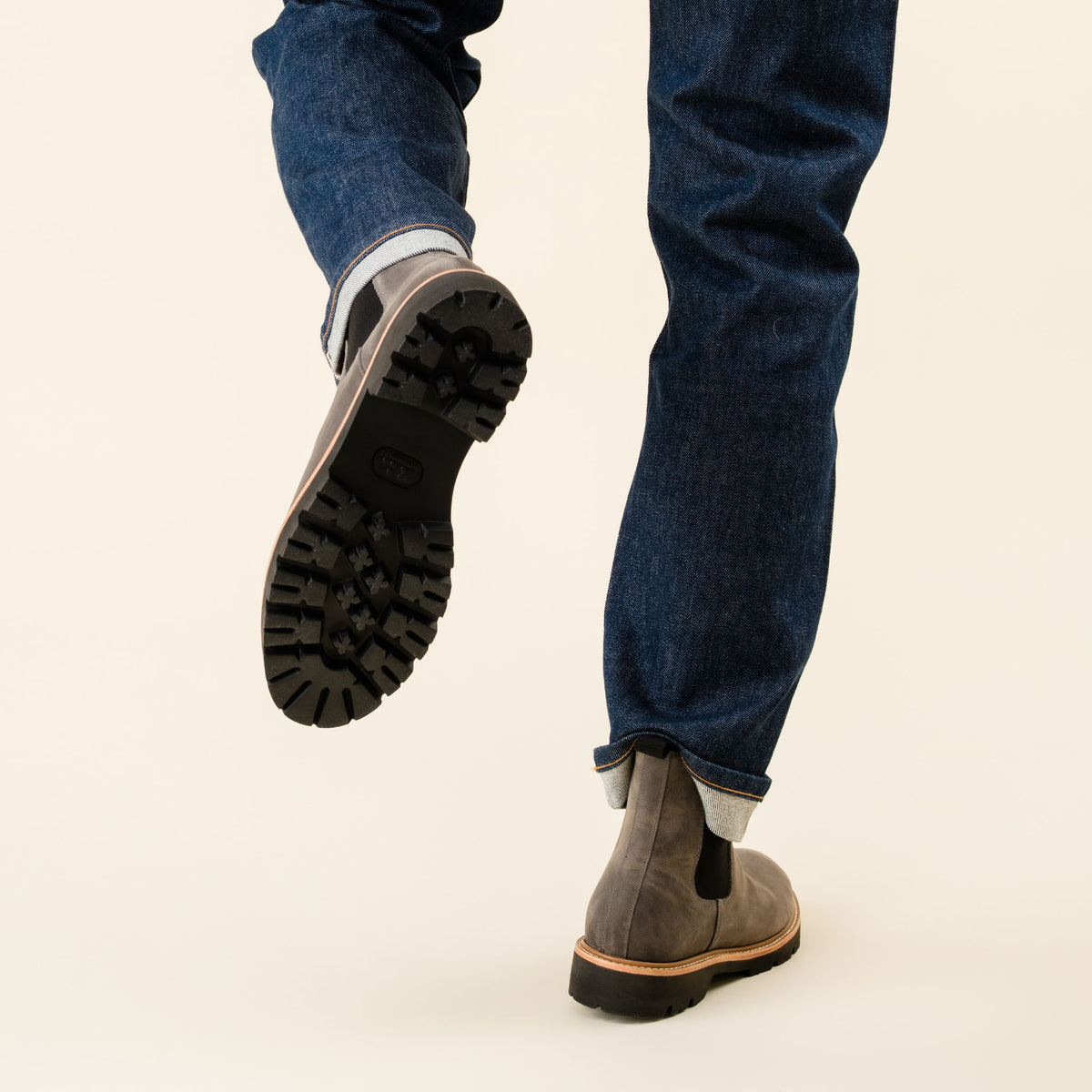 Grey leather Chelsea boots with black lug soles worn with blue denim jeans, showing the tread on one lifted foot.