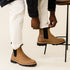 Pair of tobacco brown leather Chelsea boots with black elastic side panels and lug soles worn with white pants.