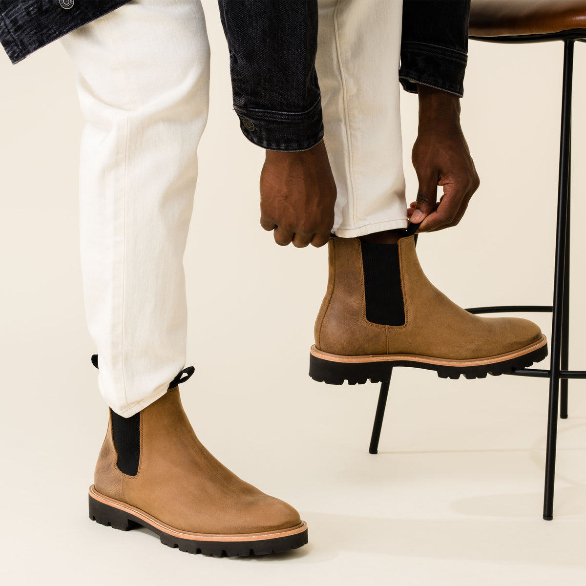 Pair of tobacco brown leather Chelsea boots with black elastic side panels and lug soles worn with white pants.