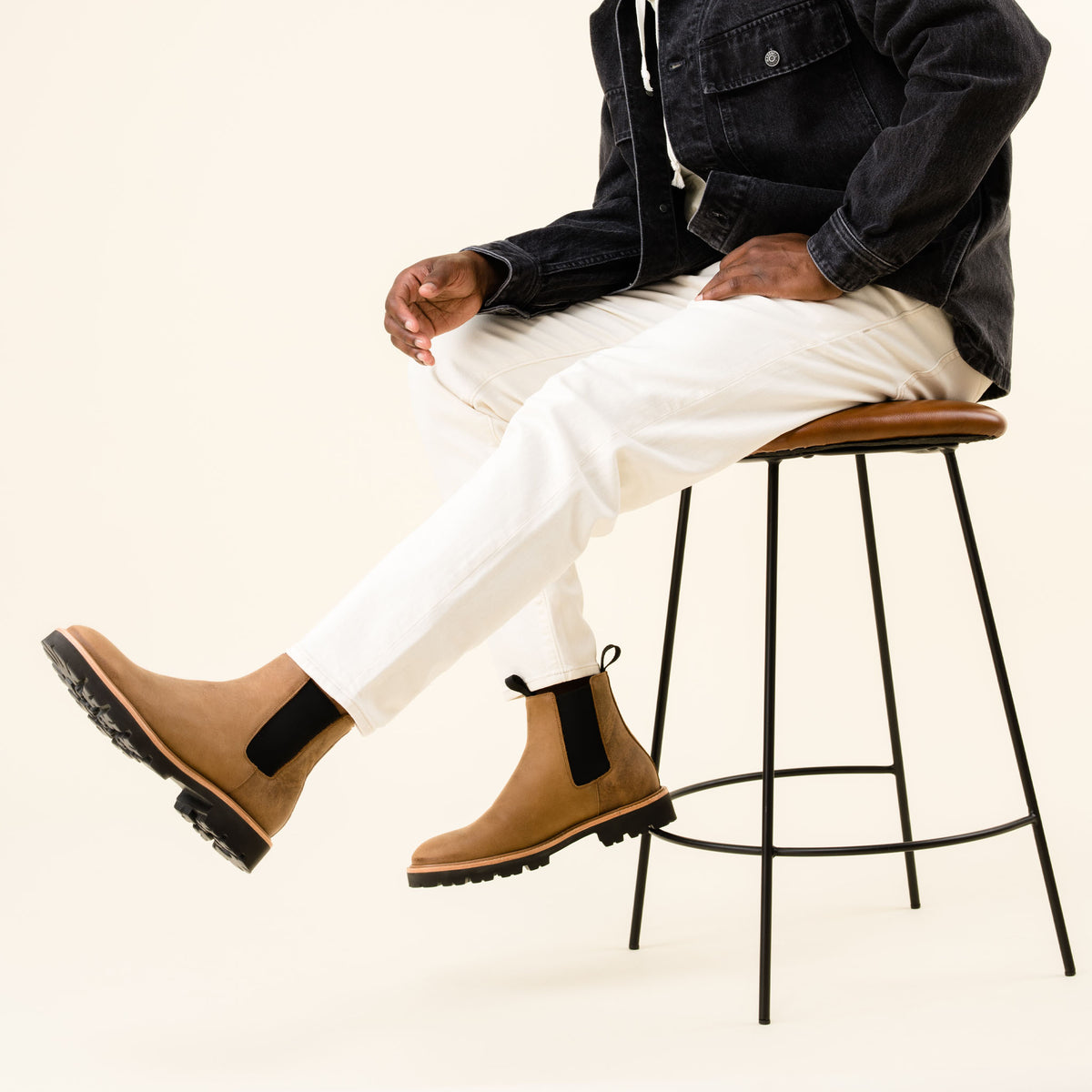 Pair of tobacco brown leather Chelsea boots with black elastic side panels worn by a person sitting on a stool.