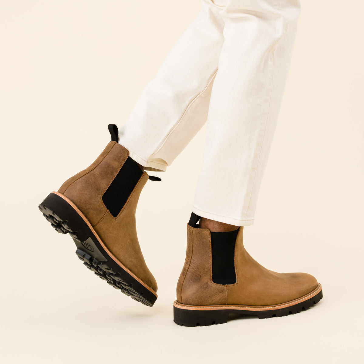 Two tobacco brown leather Chelsea boots with black elastic side panels and lug soles worn with white pants.