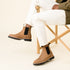 A pair of tobacco brown leather Chelsea boots with black elastic side panels and lug soles worn with white pants.