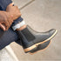 A person wearing black leather Chelsea boots with tan midsoles and black lug outsoles, paired with cuffed denim jeans.