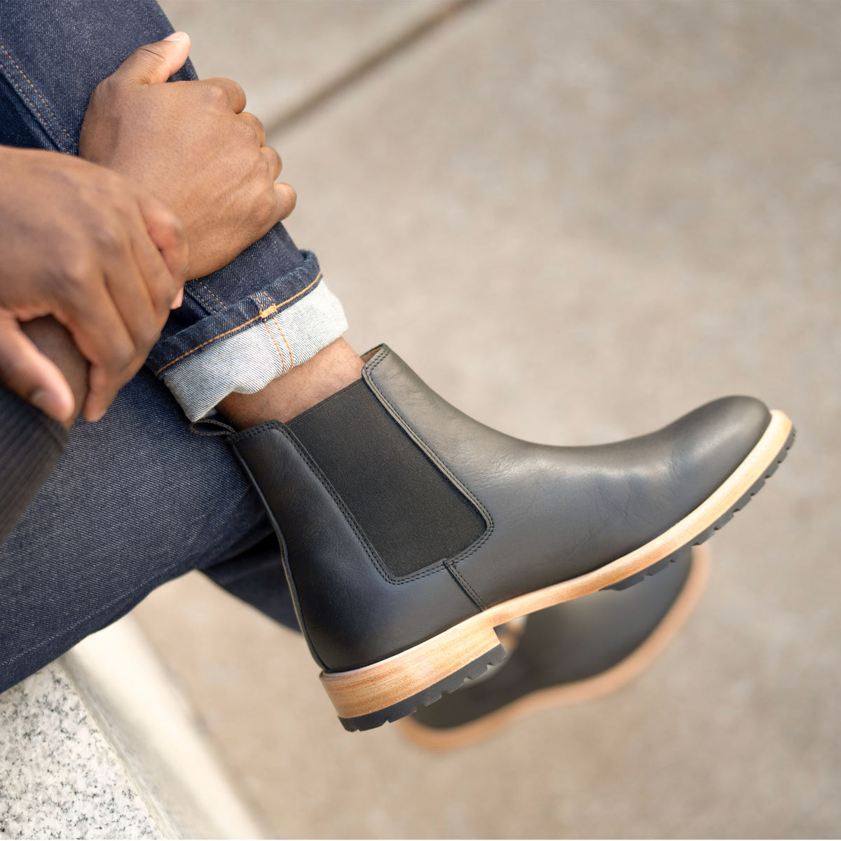 A person wearing black leather Chelsea boots with tan midsoles and black lug outsoles, paired with cuffed denim jeans.