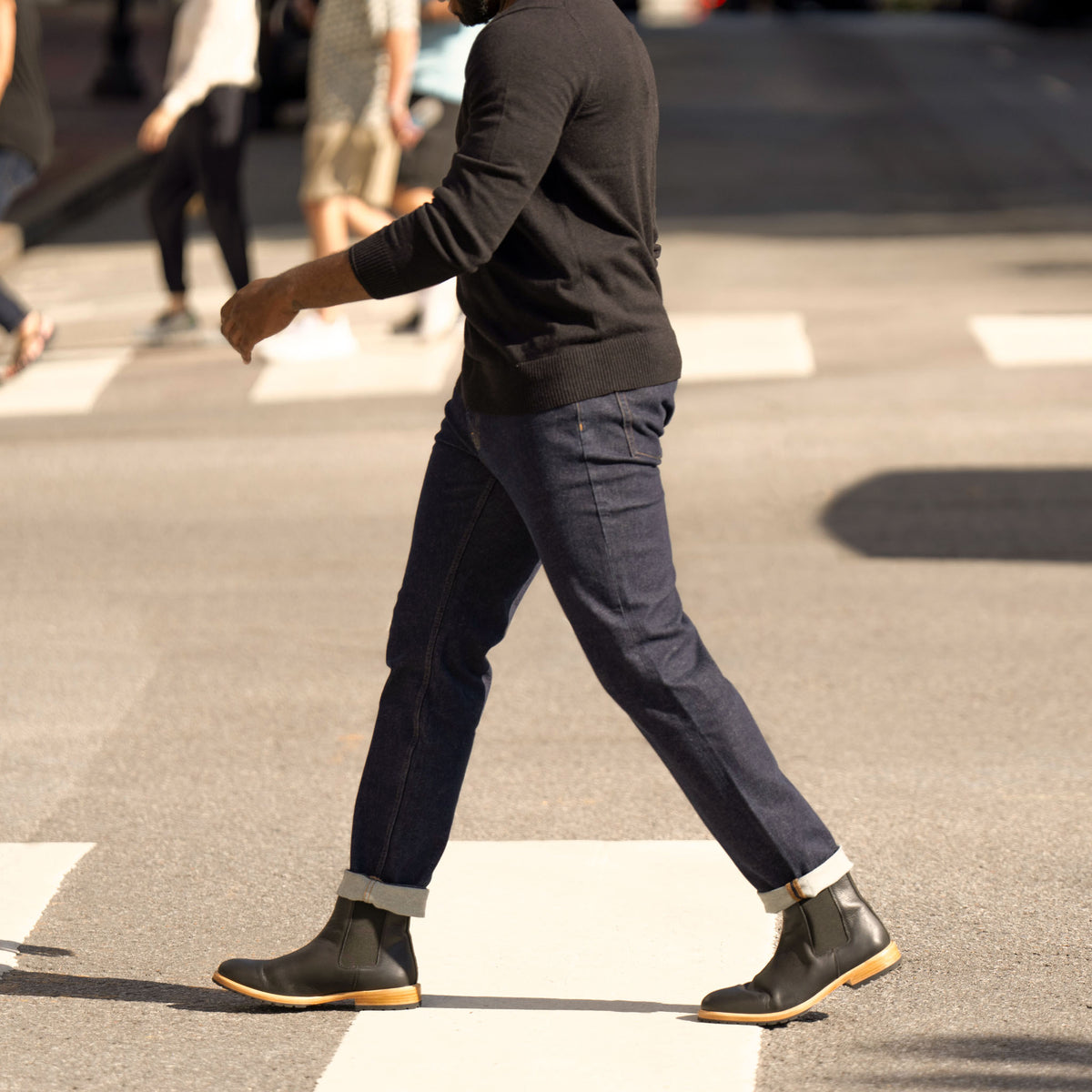 A person walking in black leather Marco Everyday Chelsea Boots with tan soles, dark jeans, and a black sweater.