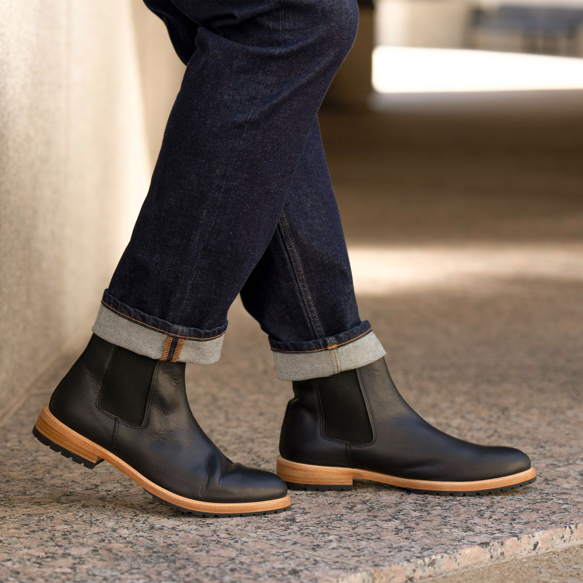 Pair of black leather Chelsea boots with tan midsoles worn with cuffed blue jeans on a stone surface.