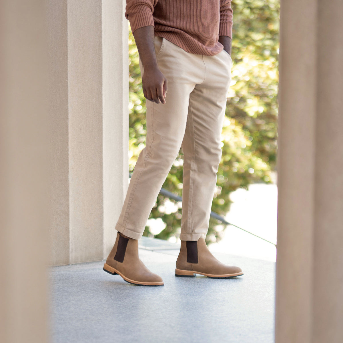 A man wears tan leather Marco Everyday Chelsea boots with dark elastic side panels, tan trousers, and a brown ribbed sweater.