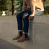 A person wearing tobacco brown leather Chelsea boots and blue jeans, sitting on a stone ledge outdoors.
