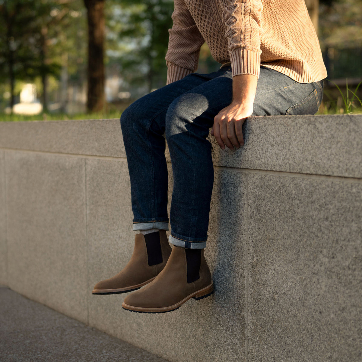 A person wearing tobacco brown leather Chelsea boots and blue jeans, sitting on a stone ledge outdoors.