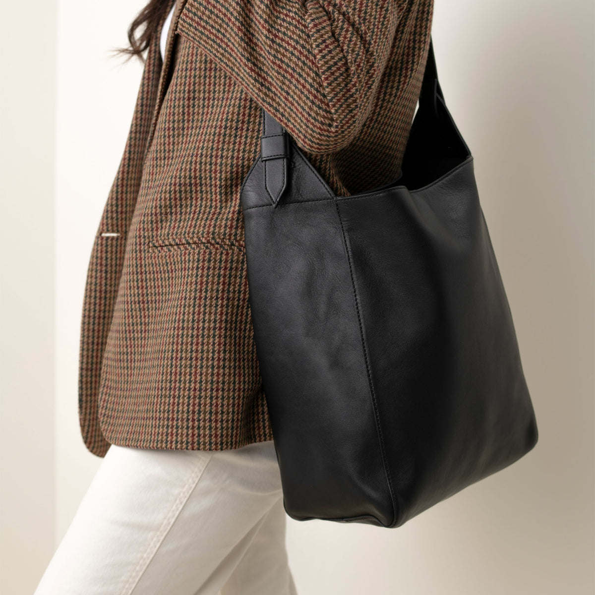Black leather Cecilia Go-To Shoulder Bag worn over the shoulder of a person in a brown patterned blazer.