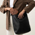 Black leather Cecilia Go-To Shoulder Bag with a brown NISOLO wallet being placed inside by a person in a plaid blazer.