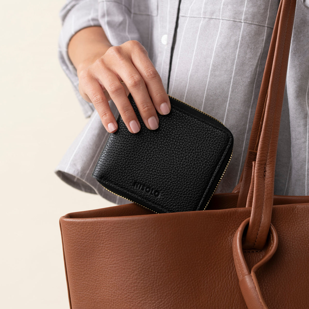 Black textured leather Remi Zip Wallet with NISOLO logo being placed into a brown tote bag.