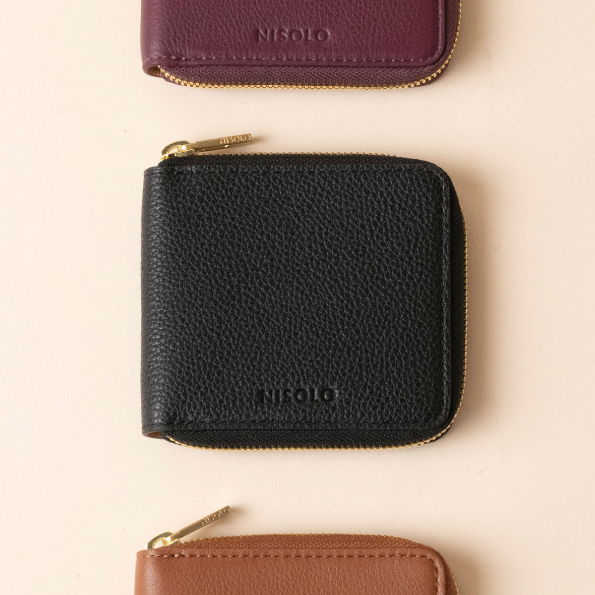 Three square leather wallets in black, plum, and brown with gold zippers and the NISOLO logo embossed on the front.